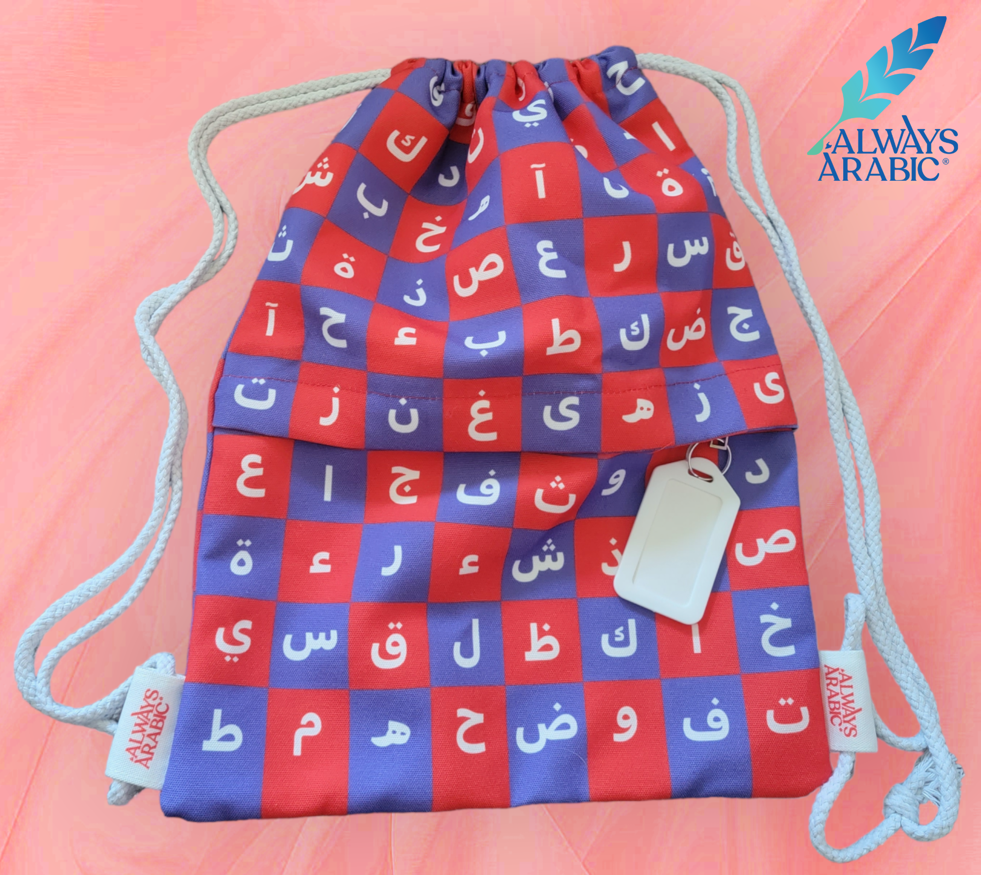 A drawstring bag with checkered blue and red fabric. Each box has a different Arabic character in it. The wordmark for Always Arabic, along with its logo (a teal feather) are displayed in the corner. 