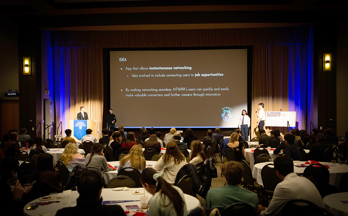 A photo of students presenting at the Driehaus Cup pitch competition. The photo includes a large PowerPoint presentation in front of a room of people sitting at tables.