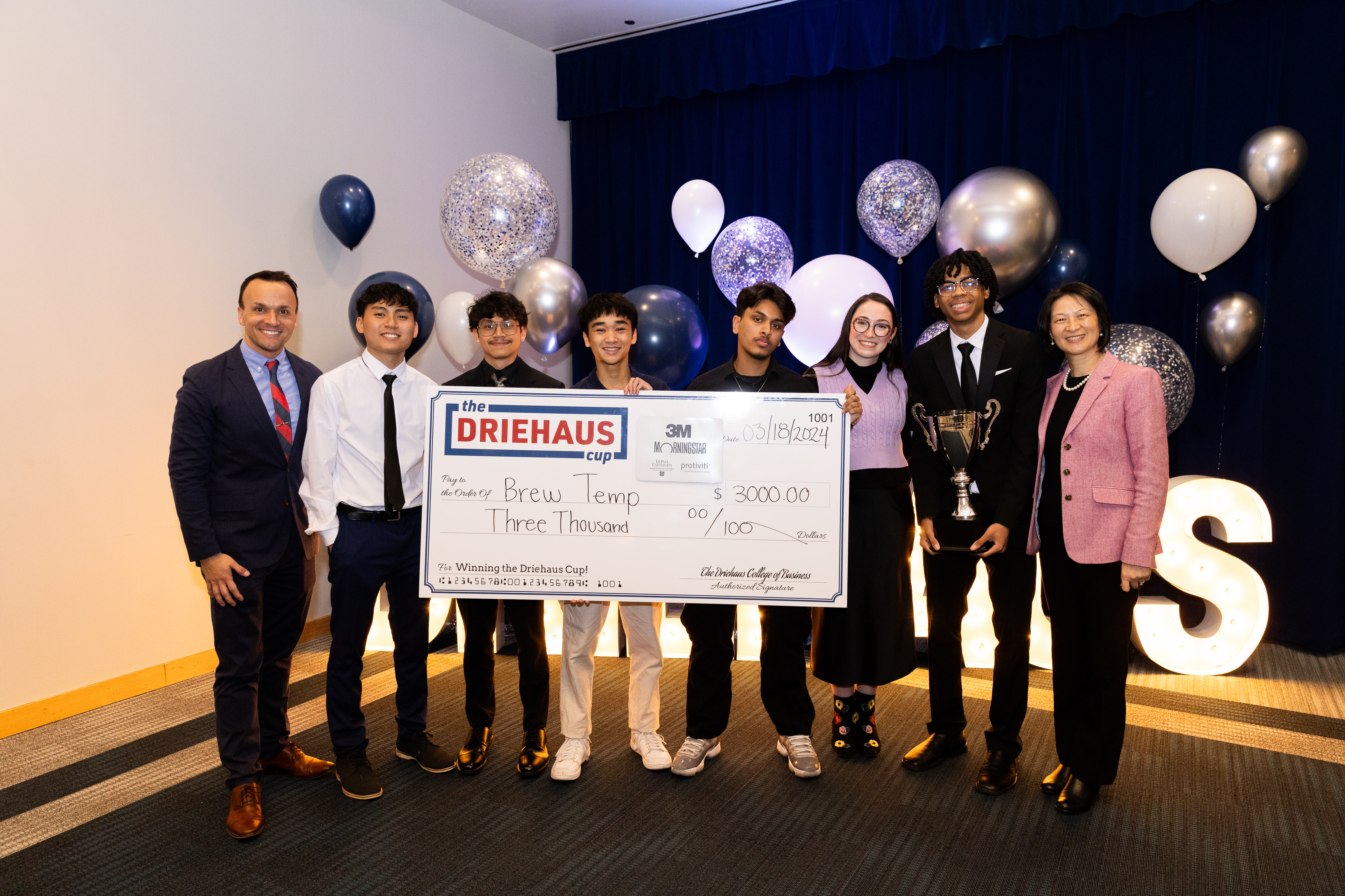 A group of college students, flanked by faculty, pose with a giant check made out to Brew Temp for $300. They are all dressed sharply, with festive yet elegant balloons behind them. 