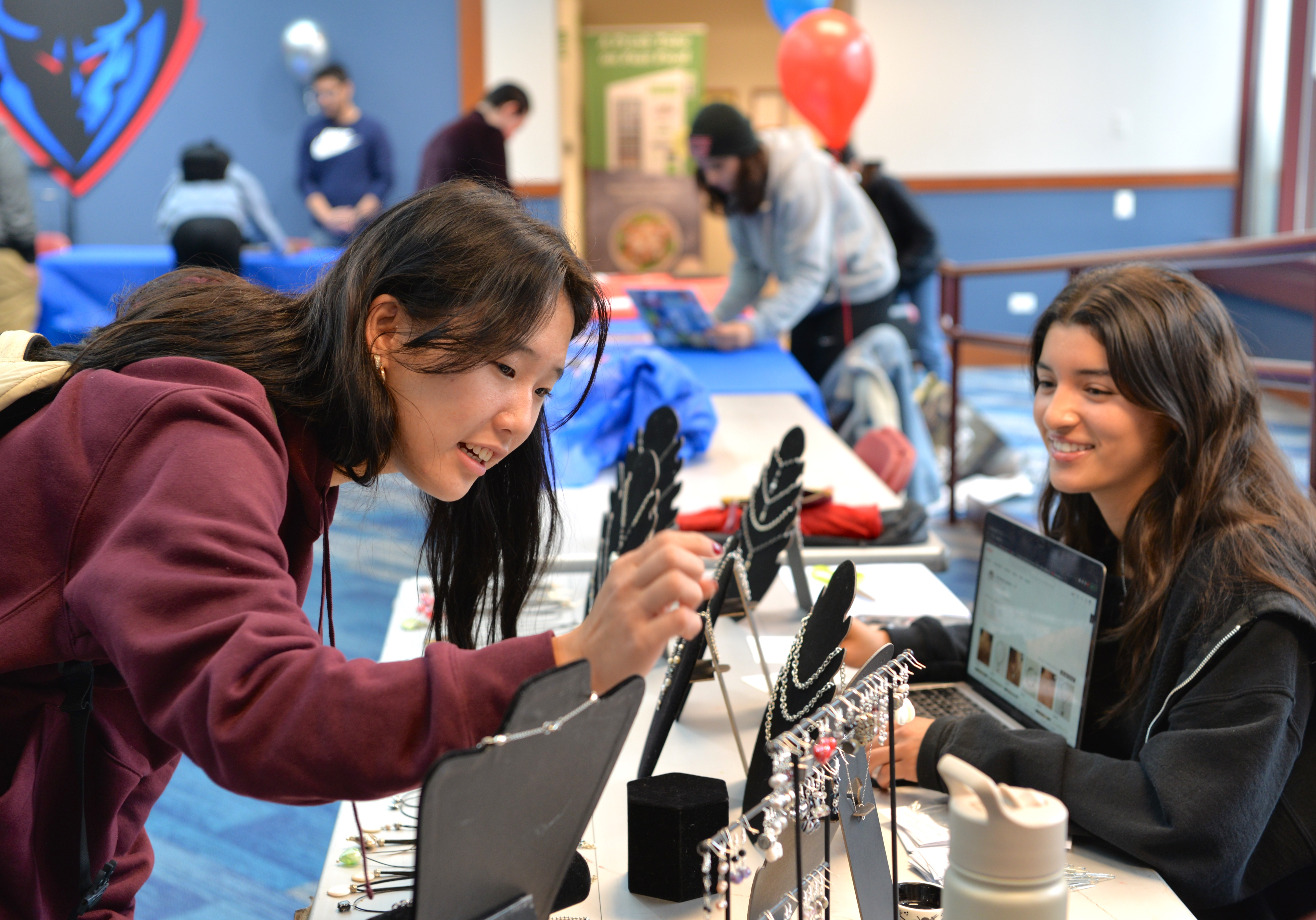 A student leans in to examine handmade jewelry displayed at a folding table, as the vendor looks on. There are brightly colored DePaul balloons in the background, as well as DePaul's vivid athletics logo, a fierce-faced depiction of a blue demon.