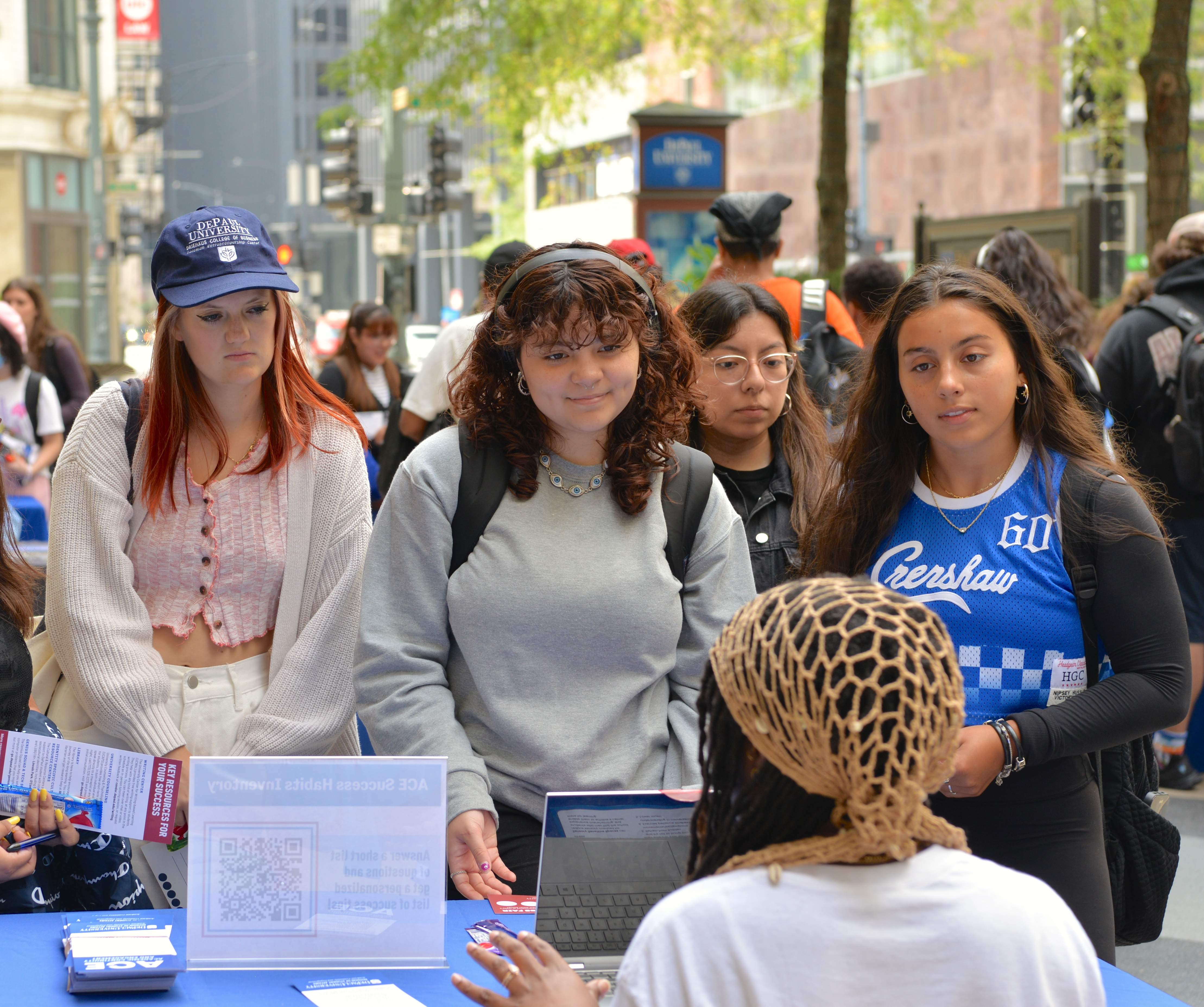 Young college students listen attentively to a person seated at a table. They are outside on a bustling city sidewalk