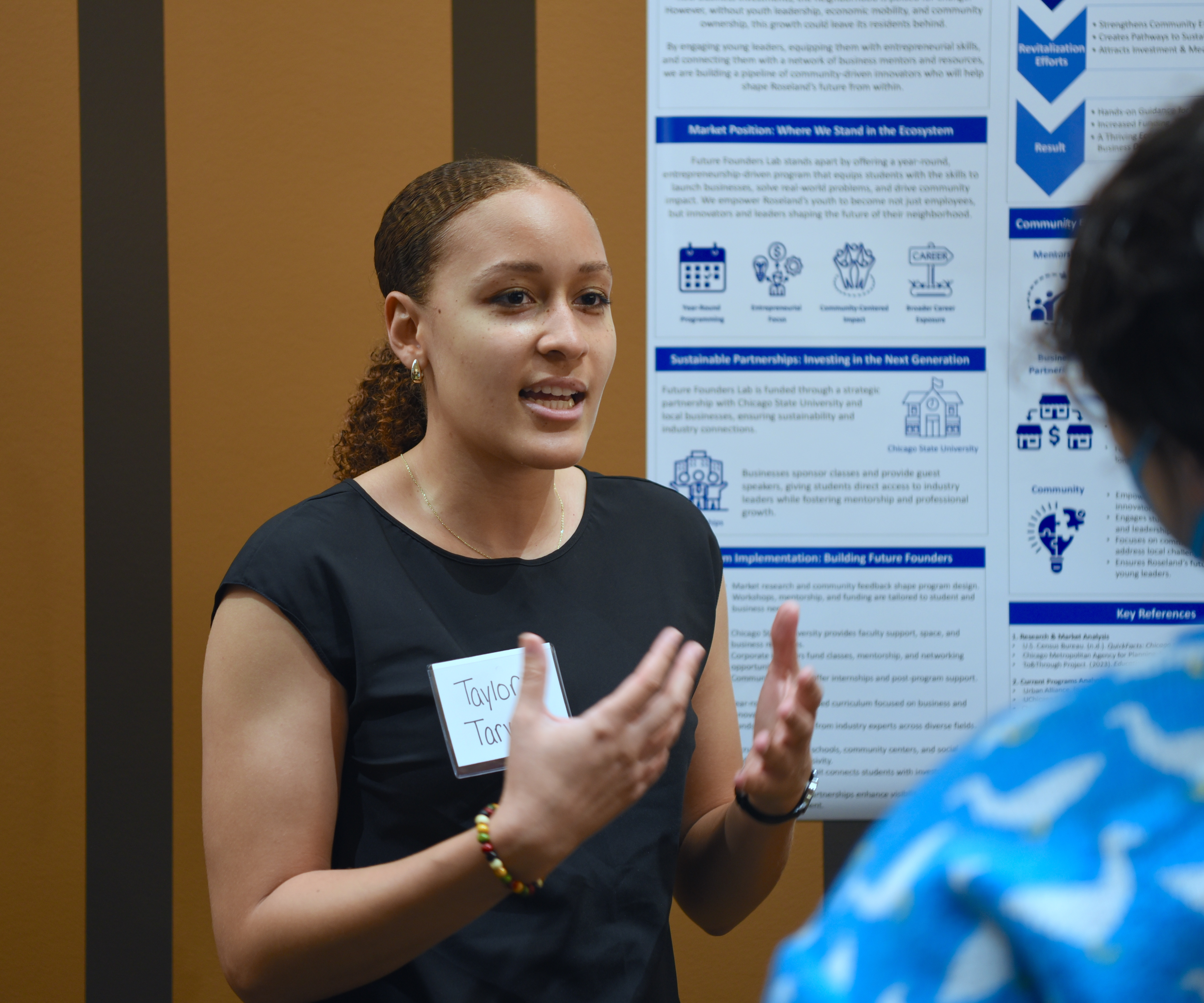 A student gestures, mid-presentation, in front of a detailed poster