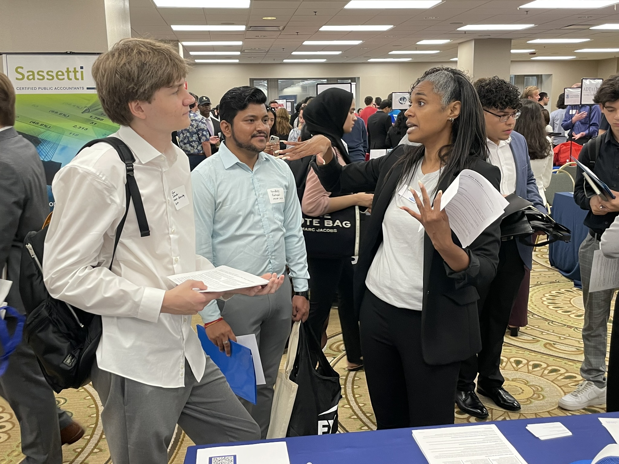 A middle-aged woman in business attire gestures as she talks to two college-age students. The setting is a hotel conference center bustling with students and companies' recruitment tables.