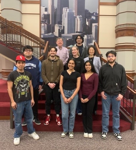 A group of students pose with two faculty in suits on a staircase, a large photo of the Chicago skyline in the background