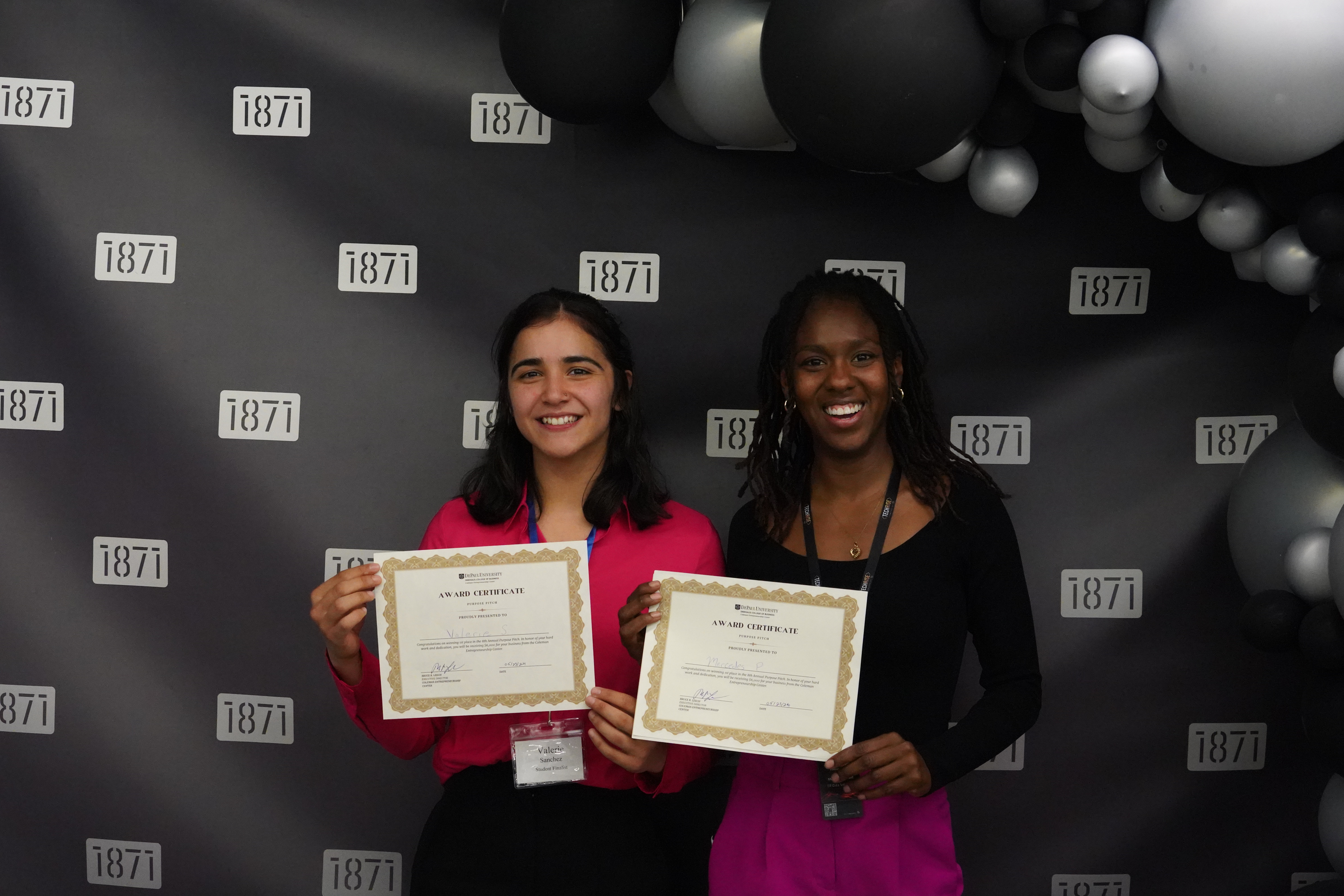 Two women hold up certificates against a black backdrop adorned with balloons