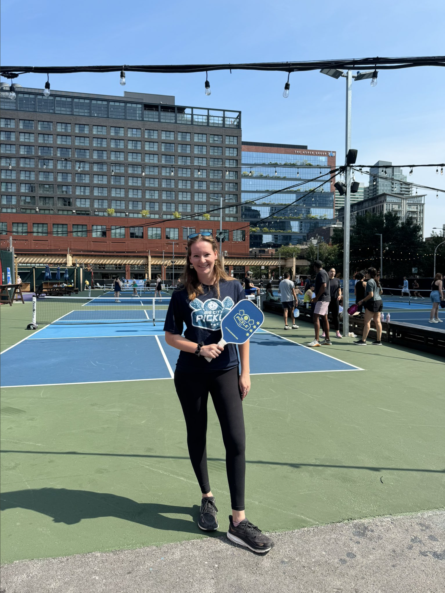 A young woman poses, racquet in hand, on a pickleball court in an urban setting