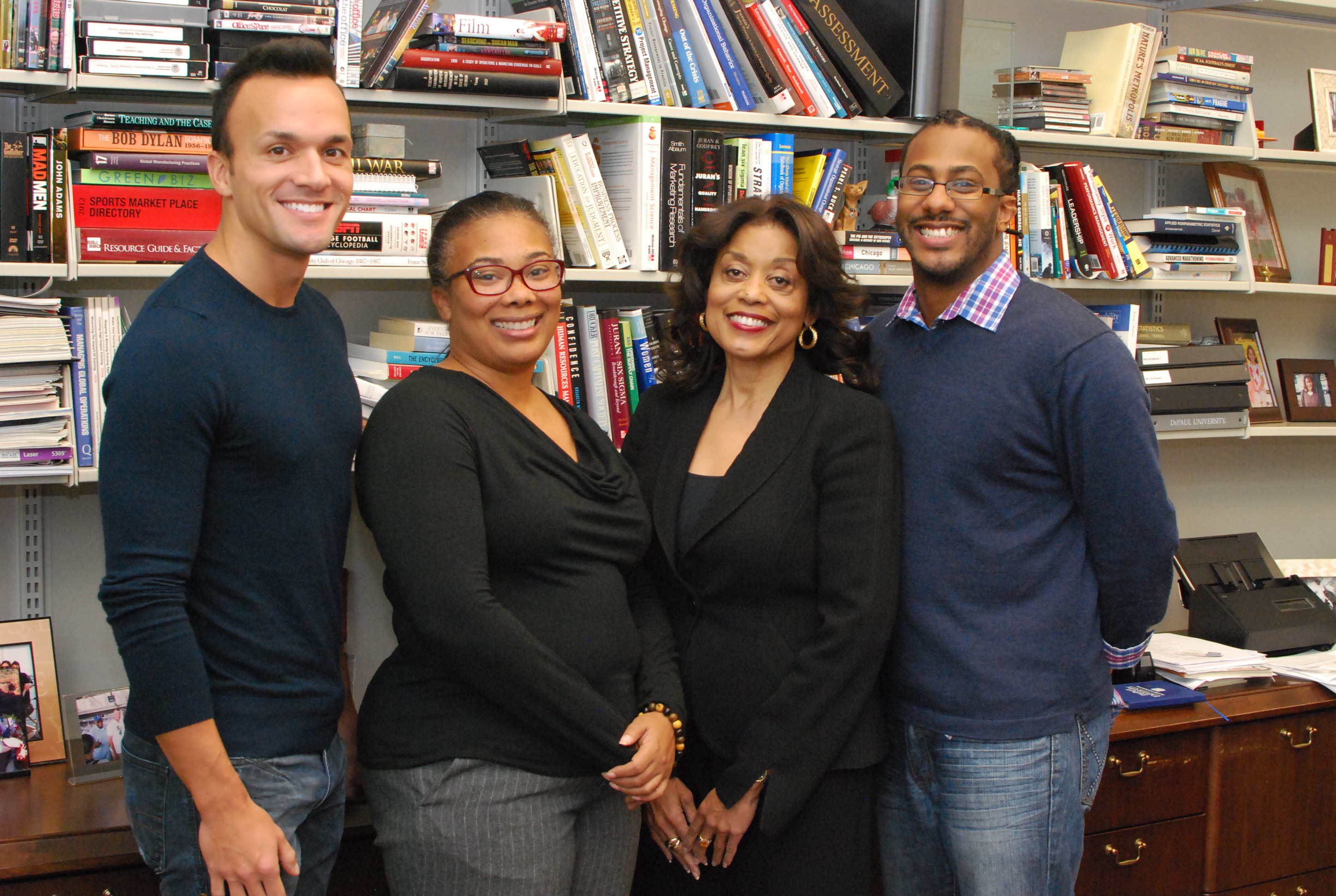 Four people in business attire pose, smiling, in front of a large bookcase