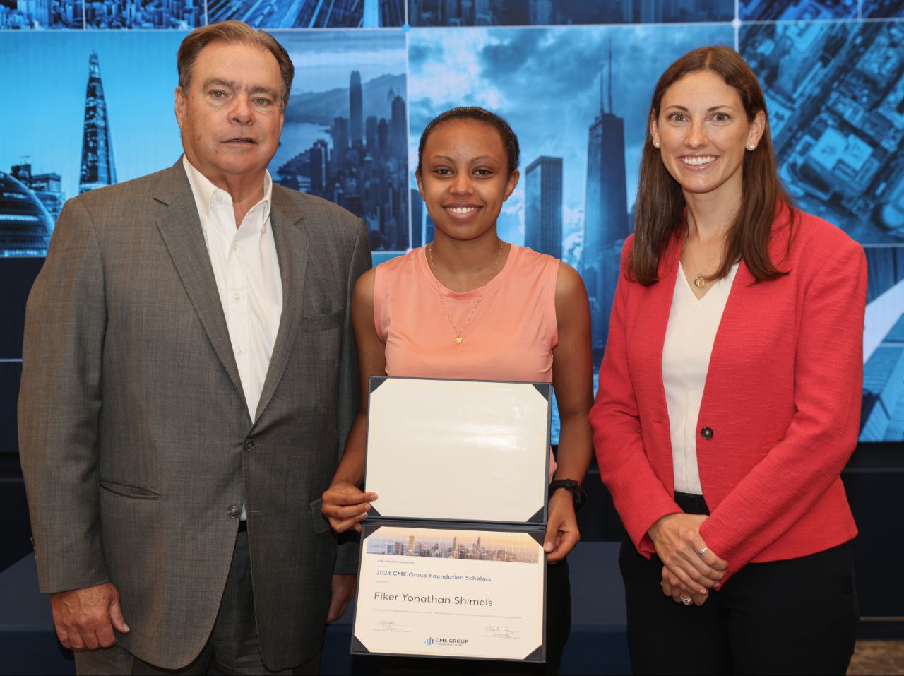 A young woman in a suit poses with a certificate in hand. Two professionals flank her and the background includes a rendering of the Chicago skyline.