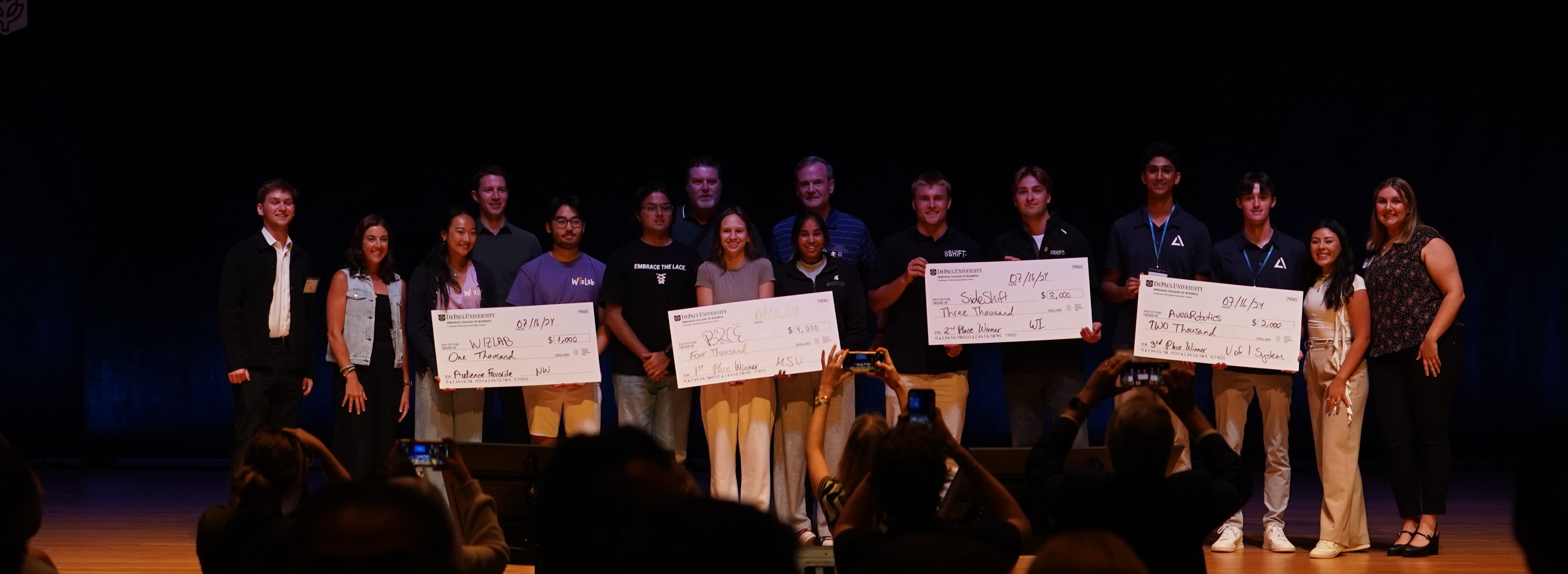 Four teams of students pose with ceremonial checks on a dimly lit stage. In the foreground, audience members can be seen taking pictures of them.