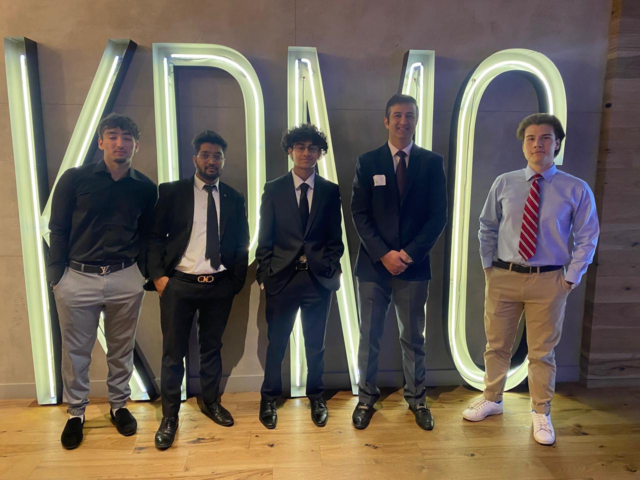 Five young men in business wear pose in front of an illuminated sign that reads KPMG