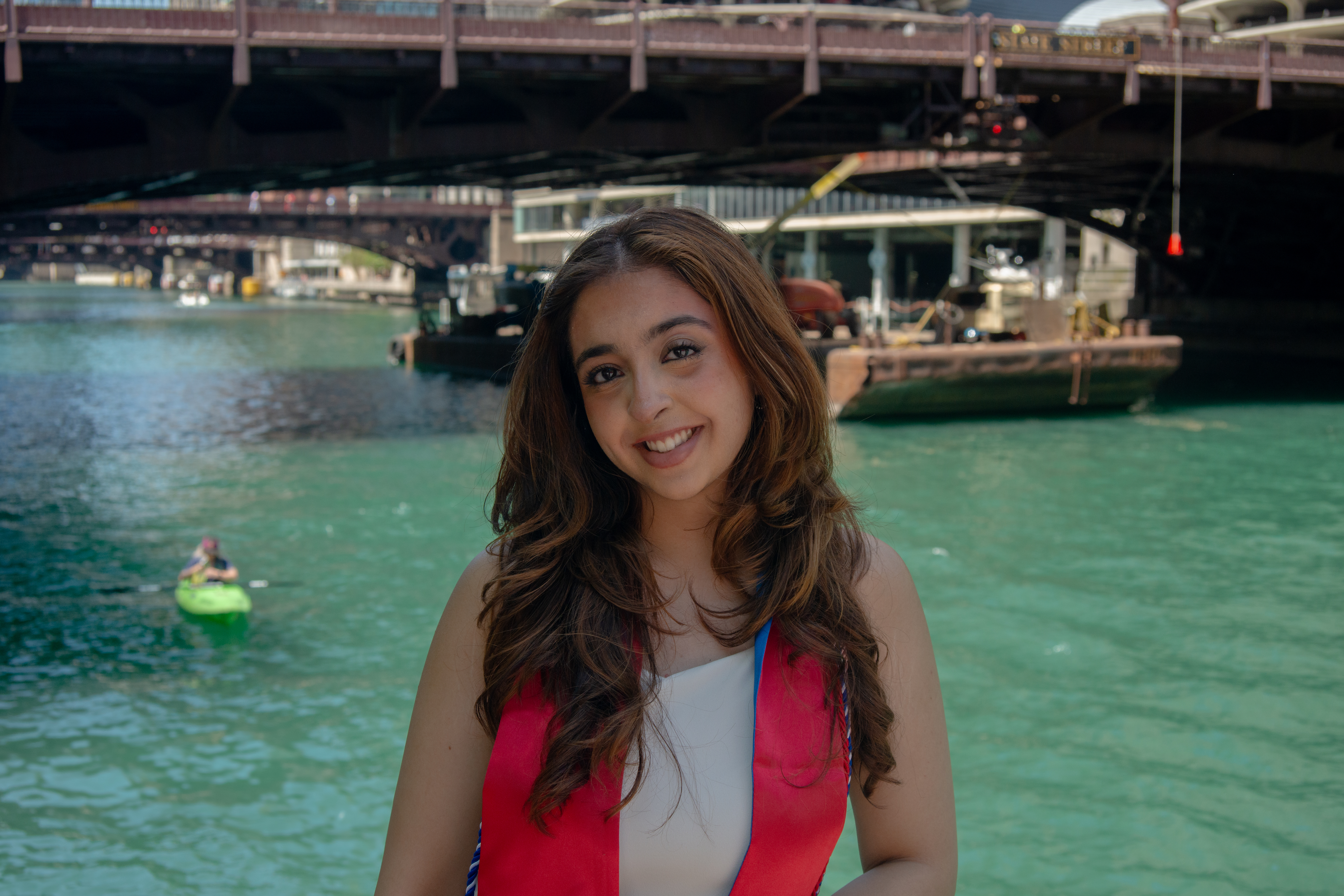 A young woman poses in front of a river with a red graduation stole