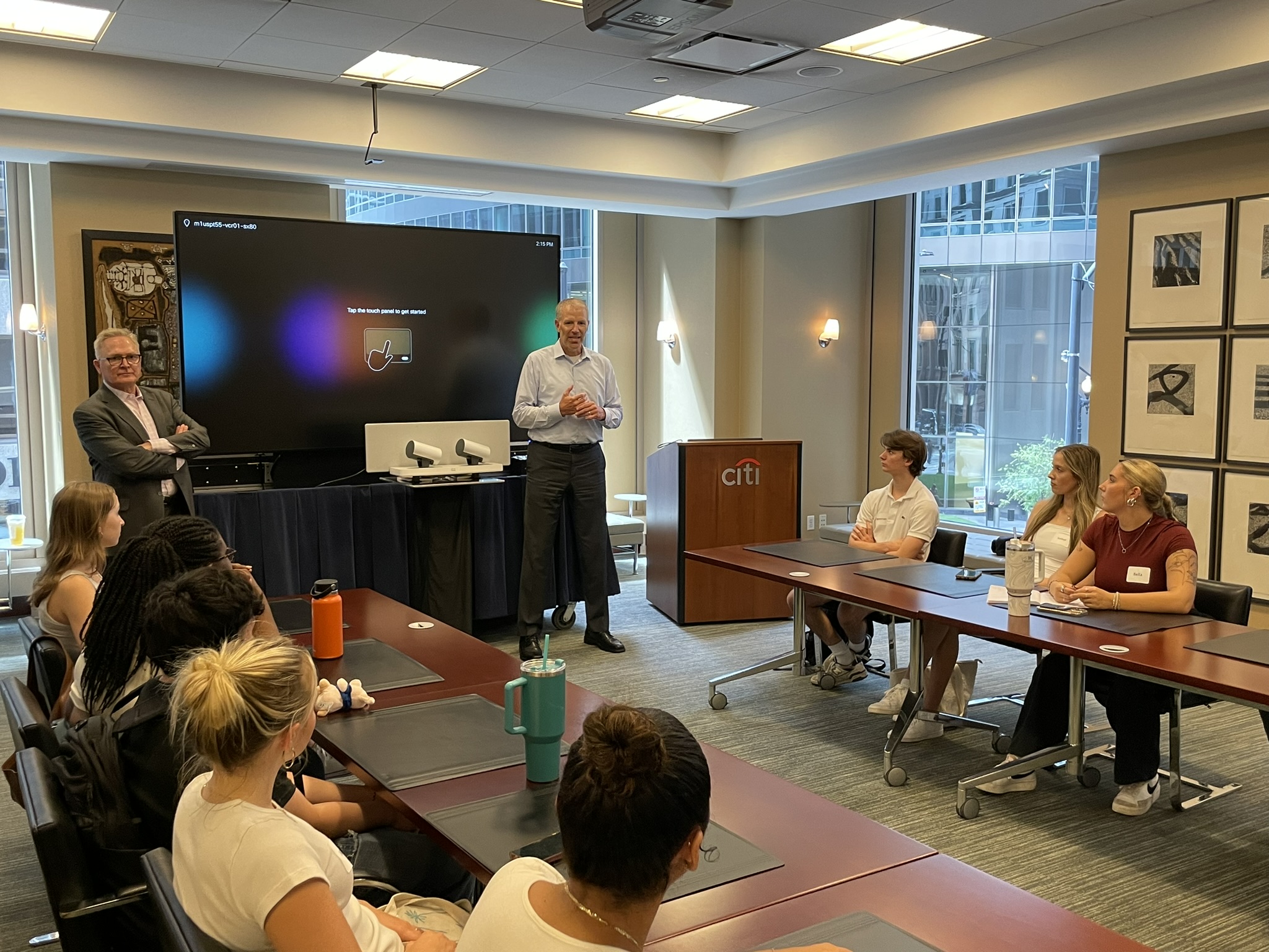 At the front of a conference room, a man in business casual attire speaks to a group of young college students. The citi logo is visible on a podium