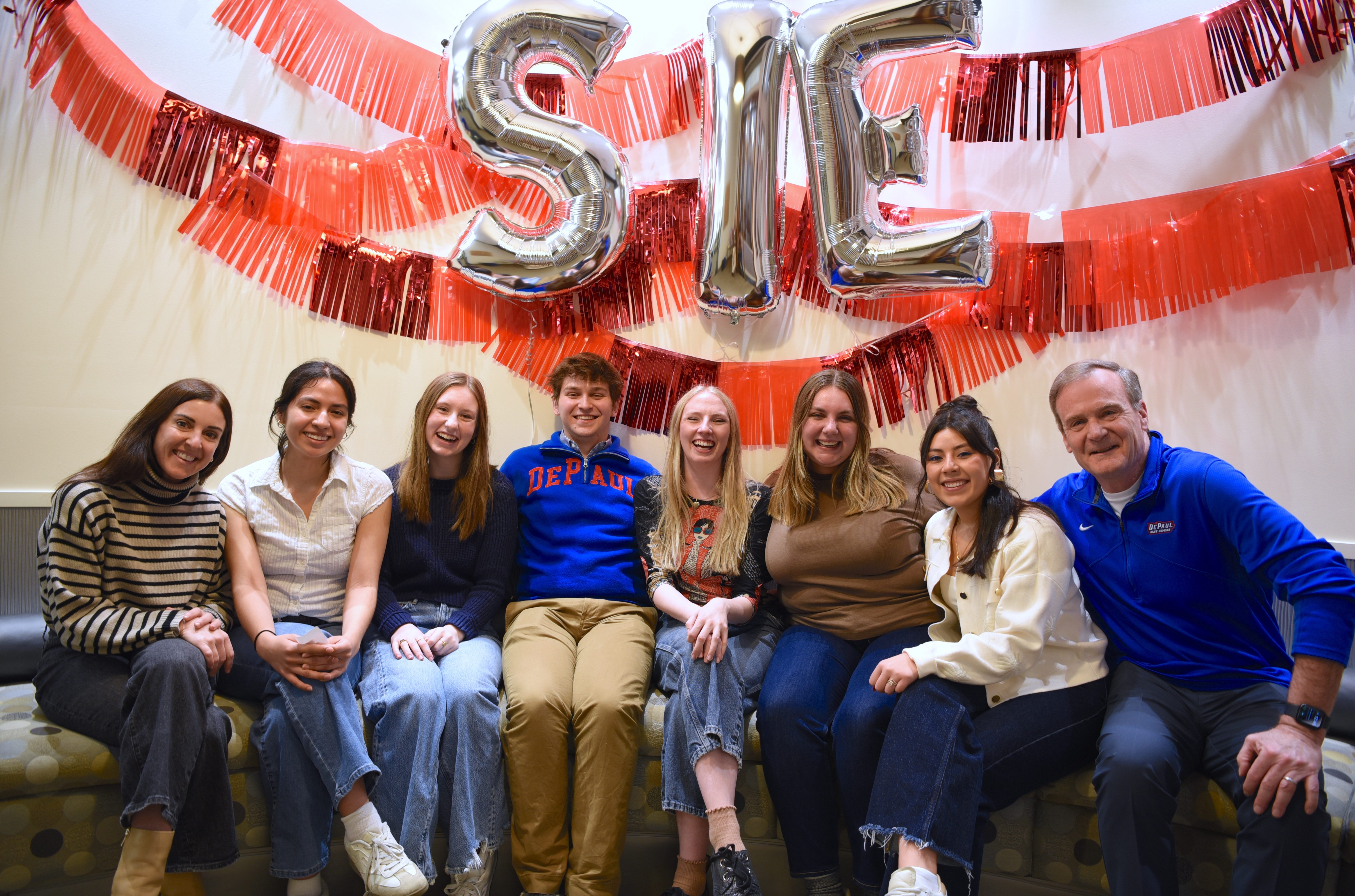 A group of faculty and students pose underneath a bright red banner and balloons spelling SIE
