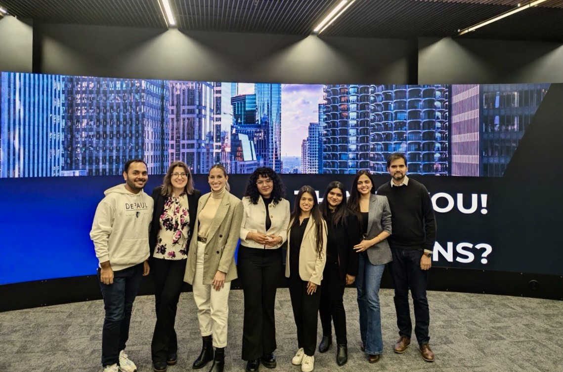 A team of eight young adults in business casual poses in front of a blue-and-purple-hued, large photo banner of iconic chicago buildings 