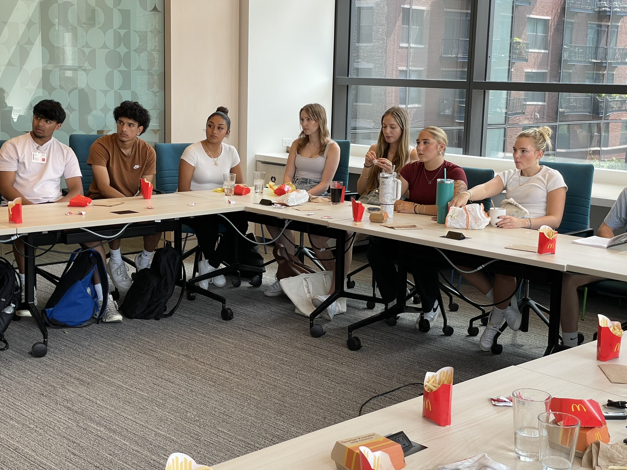 Young college students are seated at a U-shaped table in a modern conference room; they have McDonald's fries on the desks in front of them, along with laptops and notebooks