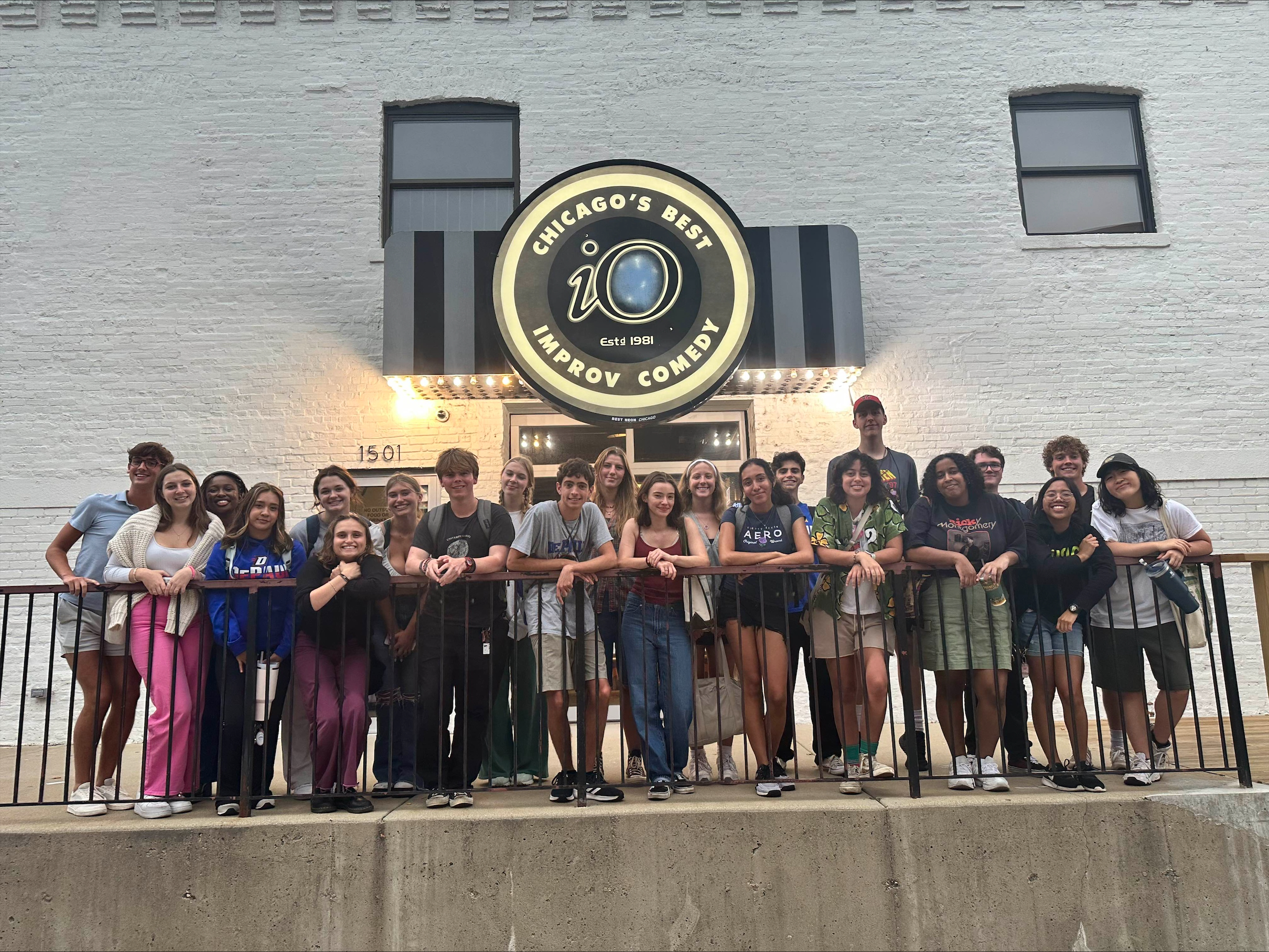 A group of students pose on a concrete balcony. On the white brick wall behind them, a prominent sign reads 