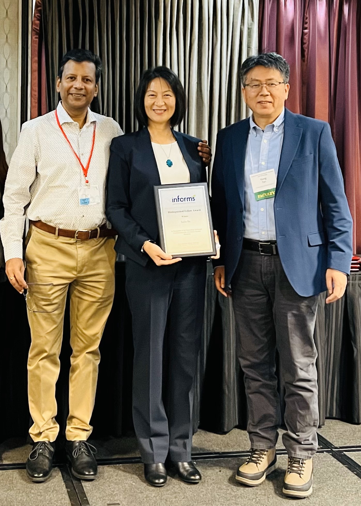 Three people in business casual wear pose in front of stage curtains. The middle person, Dean Sulin Ba, smiles as she holds a framed awards certificate