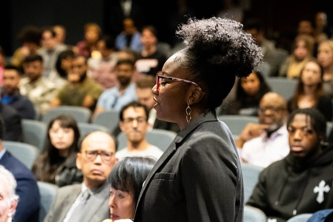 Woman in blazer standing up to speak with a full auditorium in the background